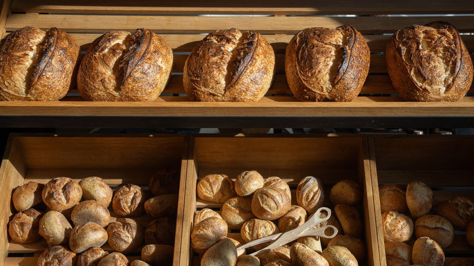 Rows of crusty sourdough loaves on a wooden shelf above crates of freshly baked bread rolls